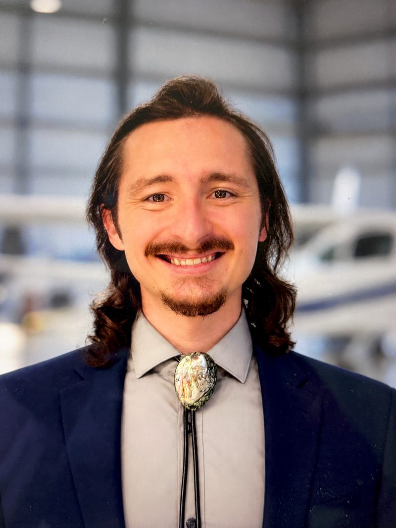 Tristen Bohn smiling in a hangar, in a navy jacket and a bolo tie, with an airplane behind him.